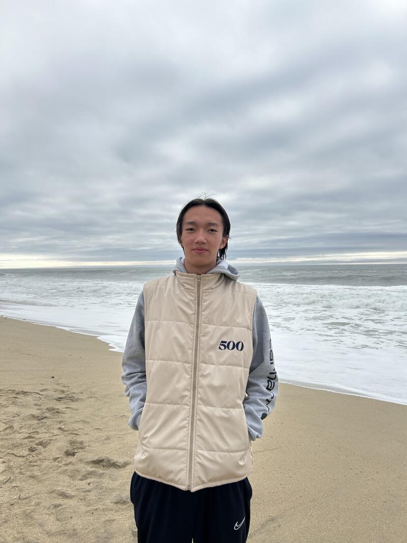 Молодой человек стоит на пляже в бежевом жилете с номером «500», в серой толстовке и черных брюках, на фоне океана и облачного неба.