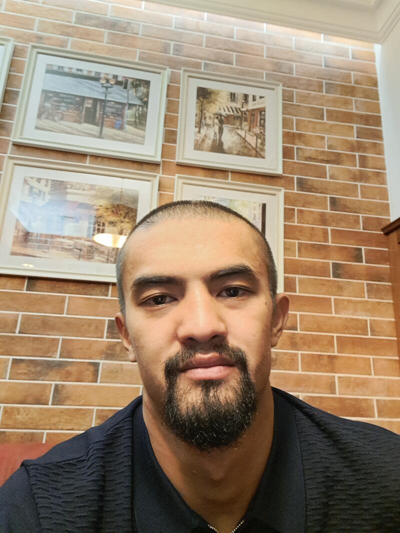 A man with a beard is posing in front of a brick wall decorated with four framed pictures.