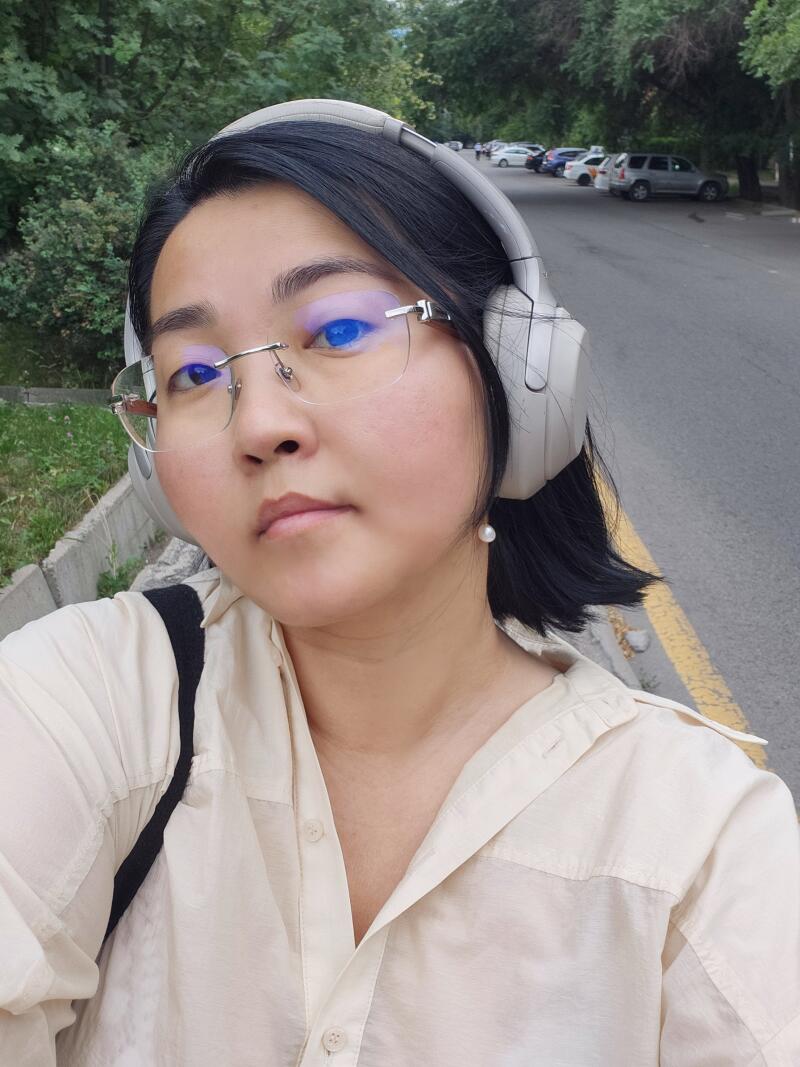 A woman with short black hair is wearing glasses and headphones, standing outdoors in a light-colored button-down shirt.