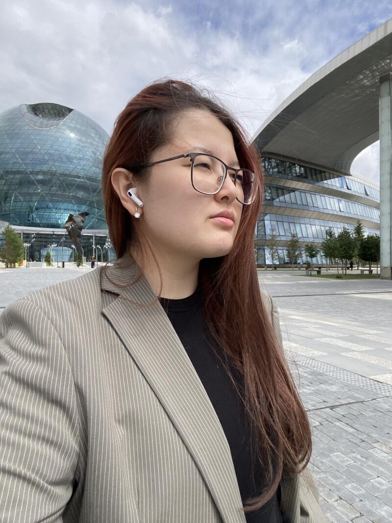 A woman wearing glasses and AirPods stands outside against a backdrop of modern architecture.