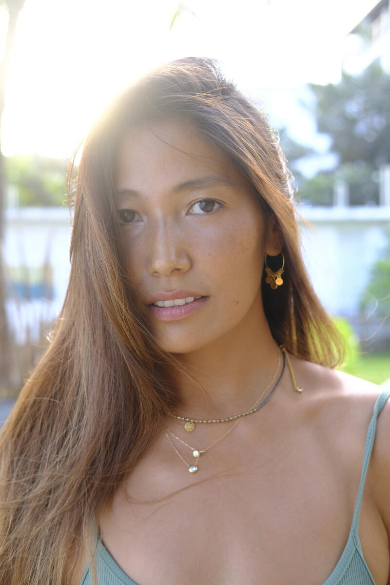A woman with long brown hair wears gold dangling earrings and layered necklaces, looking at the camera.