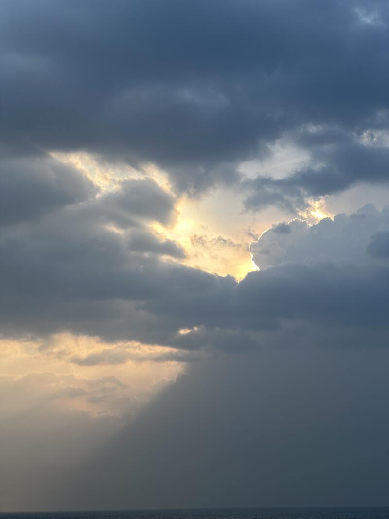 The image shows a sky filled with dark, heavy clouds, partially obscuring the sun. Sunlight streams through openings in the clouds, creating a dramatic effect.