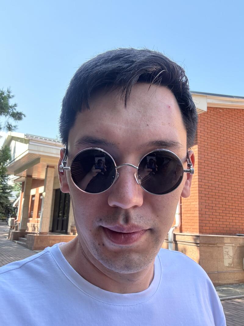 A person wearing round sunglasses and a white t-shirt is posing for a selfie outdoors, with buildings and trees visible in the background.