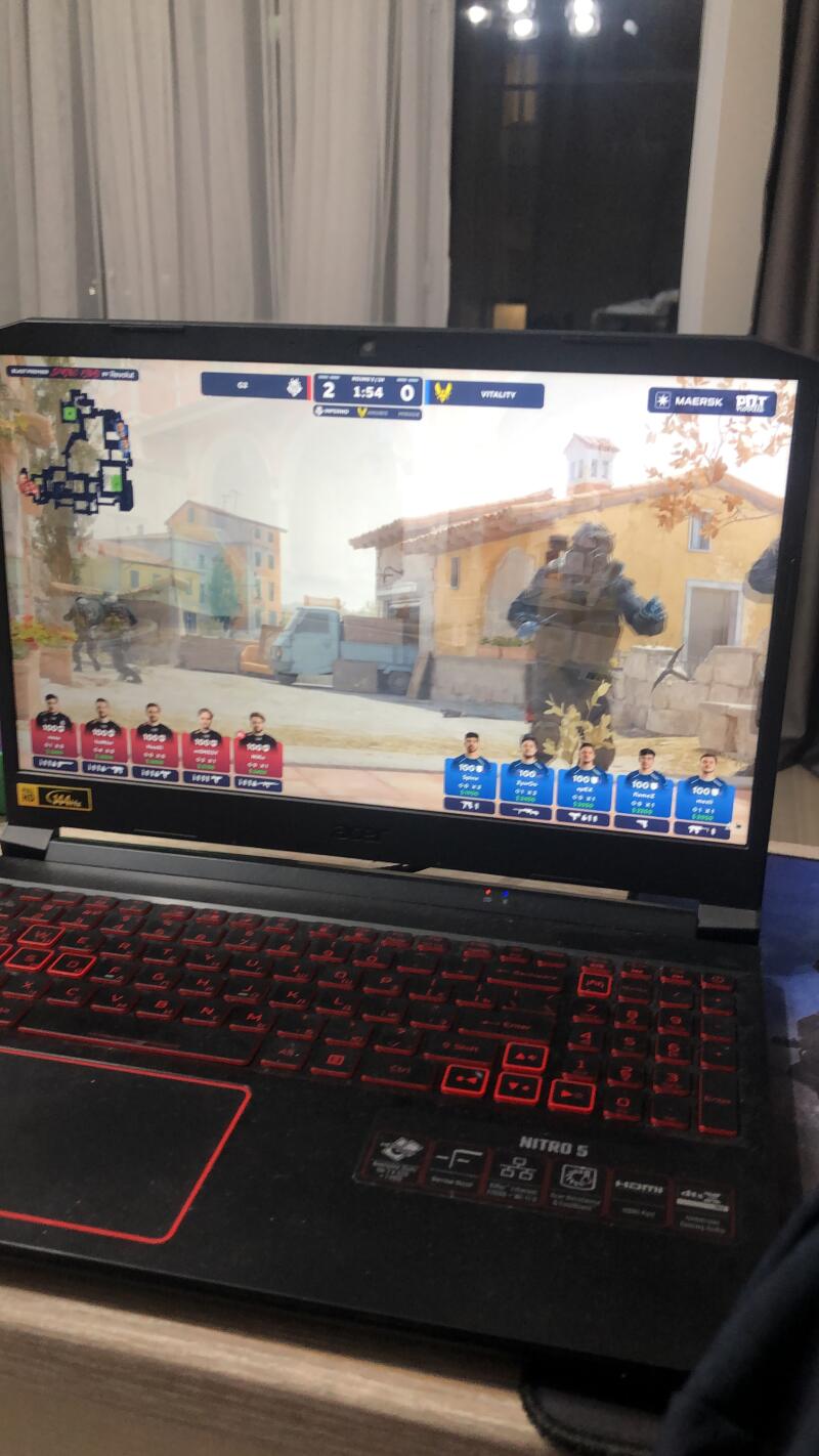 A laptop displays a Counter-Strike: Global Offensive match, showing the in-game action and the score 2-0. The laptop is an Acer Nitro 5 with a red-backlit keyboard.