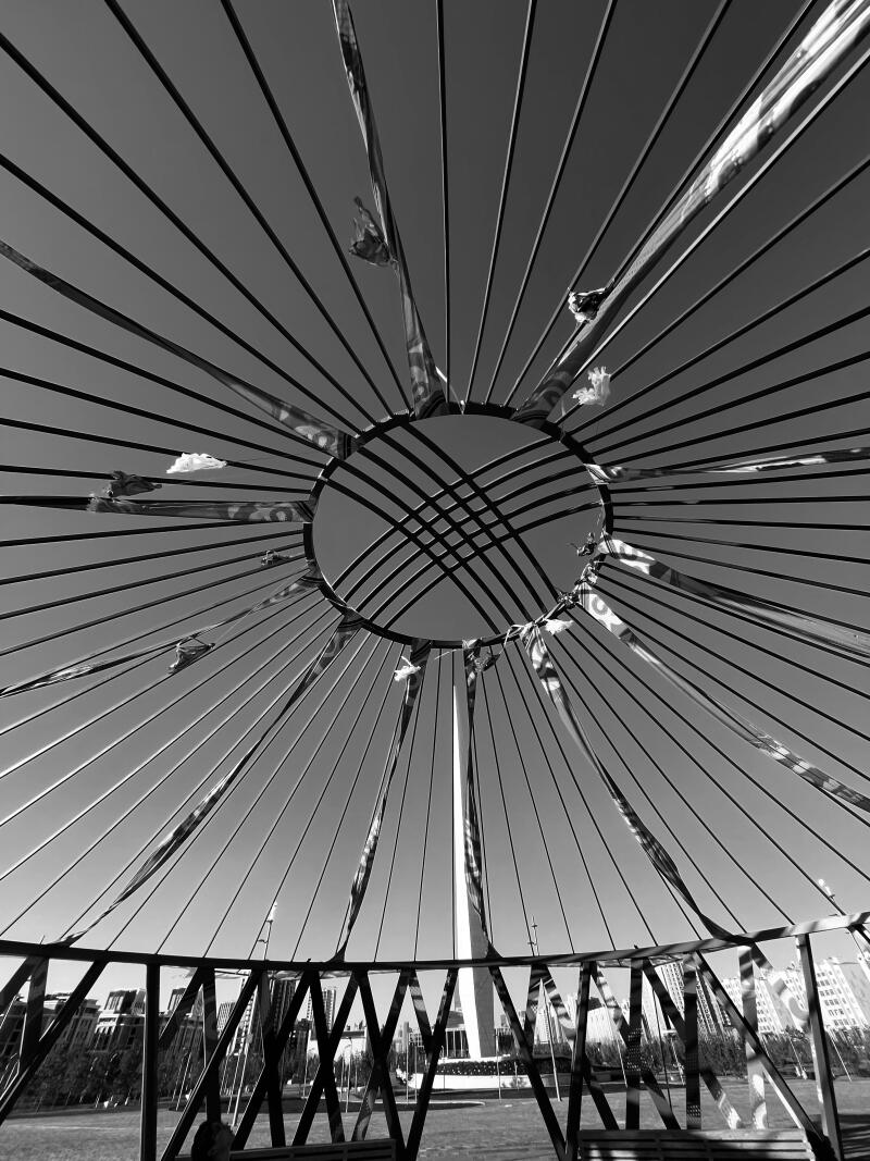 The black and white shot looks up from inside a yurt-like structure, featuring a circular opening at the top and radiating beams, with ribbons attached. In the background, a tall monument is visible.