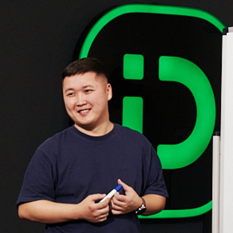 A smiling Asian man wearing a dark blue t-shirt holds a marker in his hands, standing in front of a black background with a bright green logo and a whiteboard.