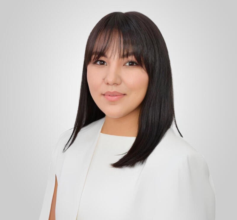 A woman with dark hair and bangs wears a white dress and jacket against a gradient gray background.