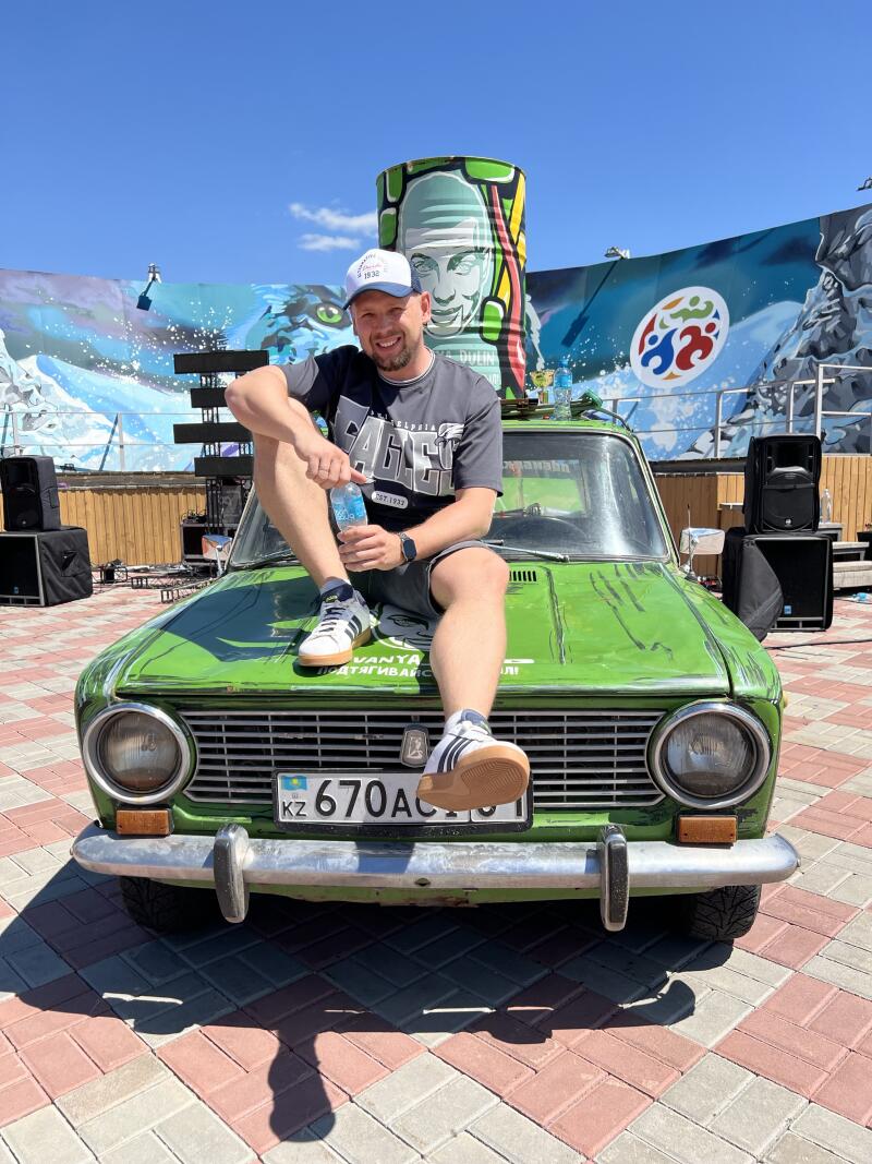 Мужчина сидит на капоте зеленой машины с номерным знаком KZ 670ACJ01, держит бутылку с водой, на фоне нарисованные фрески и колонки.