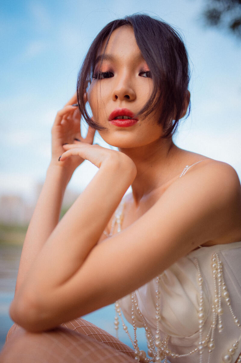 A woman with red lipstick and dark hair poses in a white top adorned with pearls, with her hands near her face.