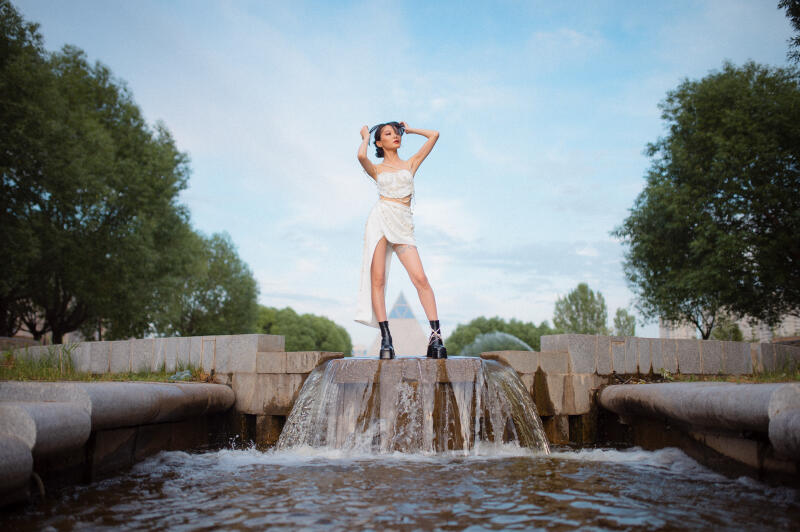 A woman in a white outfit and black boots stands on a stone fountain with water flowing around her.