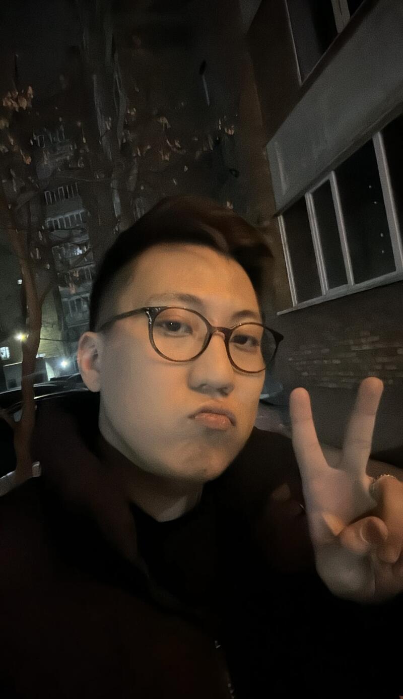 A person with glasses making a peace sign in an outdoor setting with buildings in the background.