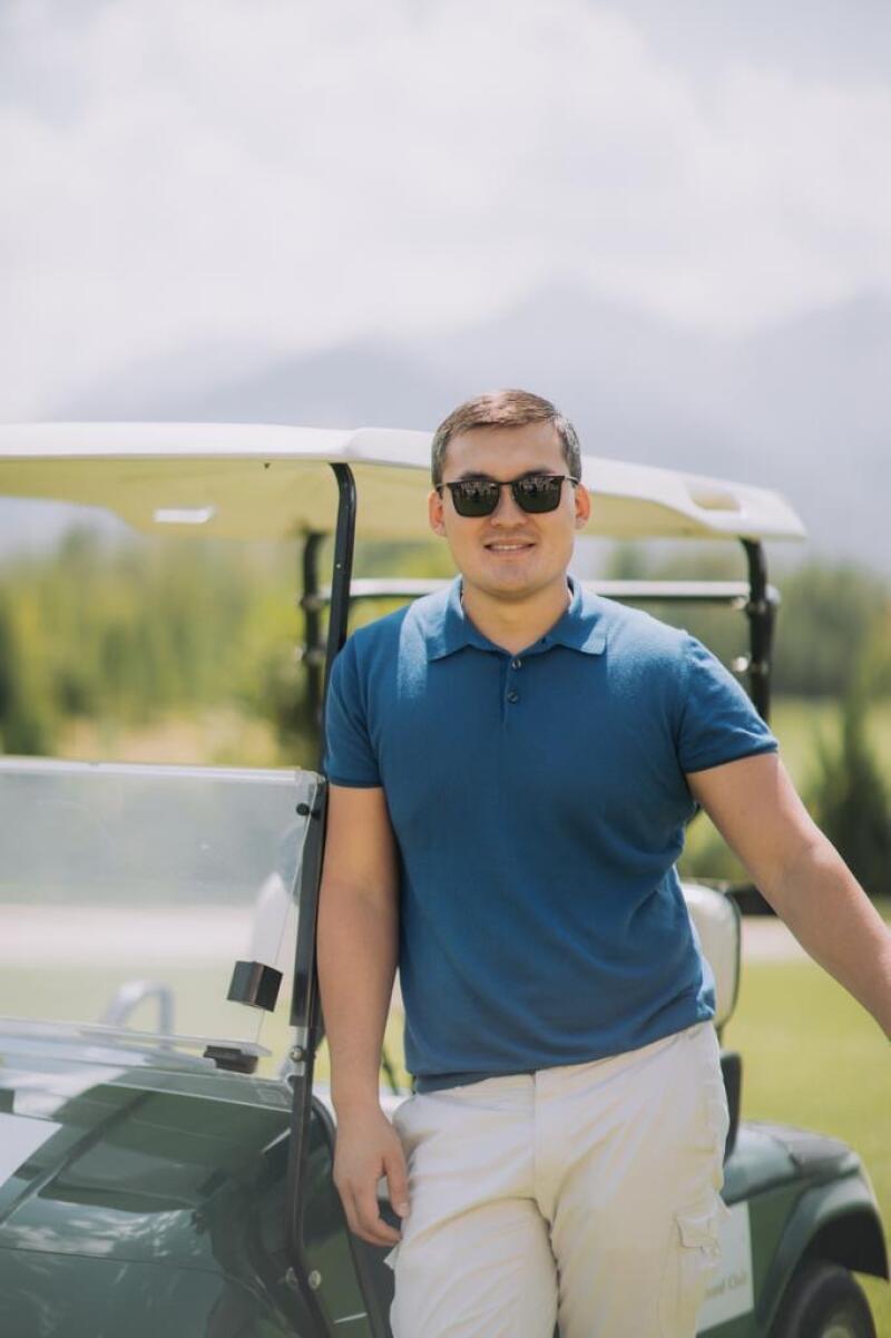 A man wearing sunglasses, a blue polo shirt, and beige pants is leaning on a golf cart in a grassy area.