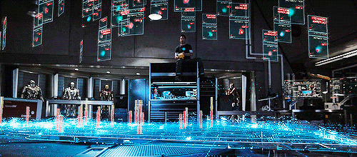 The scene shows a lab with computer screens and figures; a man sits atop a structure amidst the technological displays.