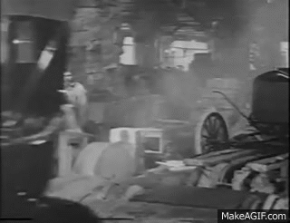 A black and white scene shows a man in a light-colored coat walking past a destroyed industrial area with debris and damaged equipment.