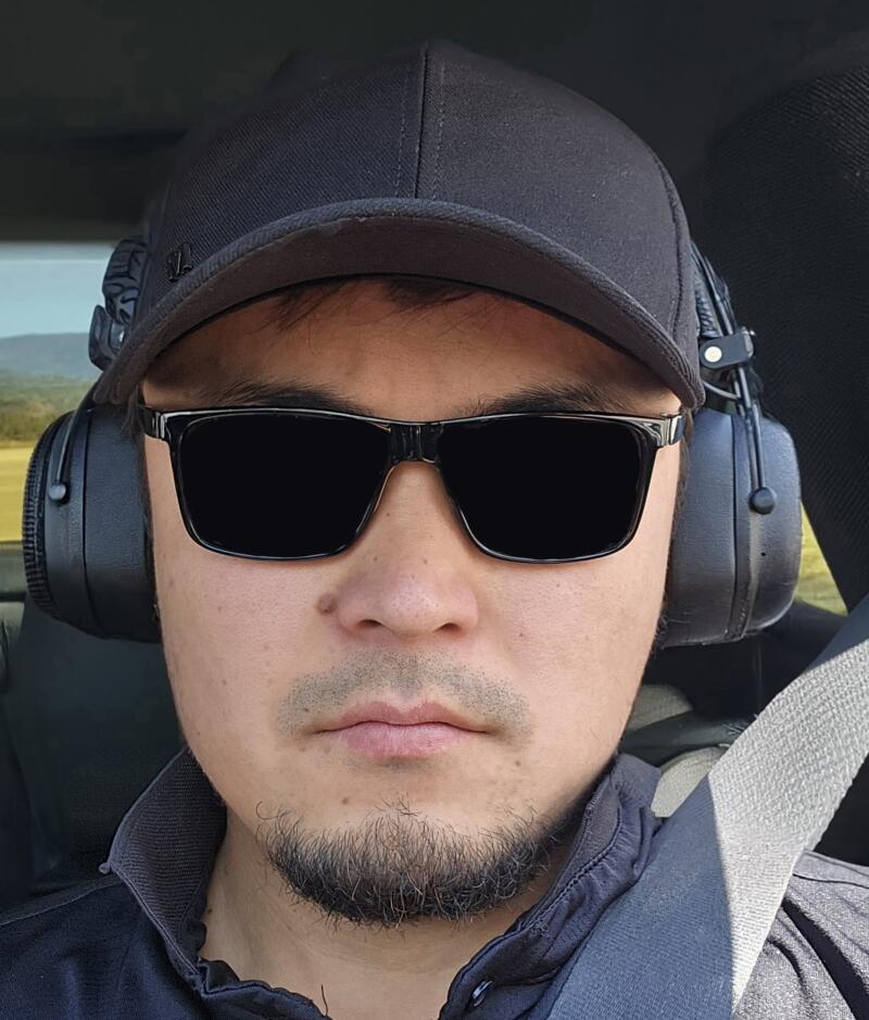A person wears a black cap, black sunglasses, and black headphones while wearing a seatbelt.