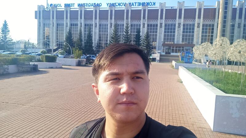 Мужчина делает селфи на фоне здания "ВОКЗАЛ КОКШЕТАУ" в солнечный день.