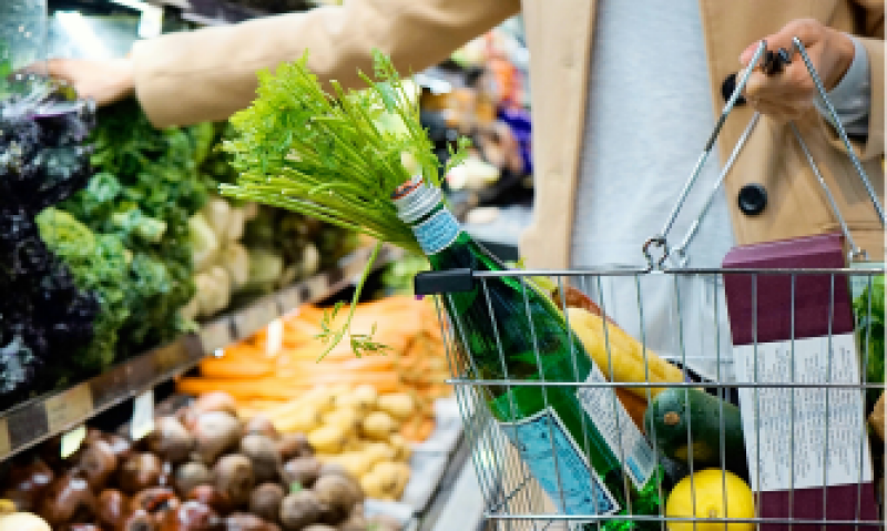 A person in a beige coat shops for groceries, filling a metal basket with items like carrots, a green bottle of water, and other produce.