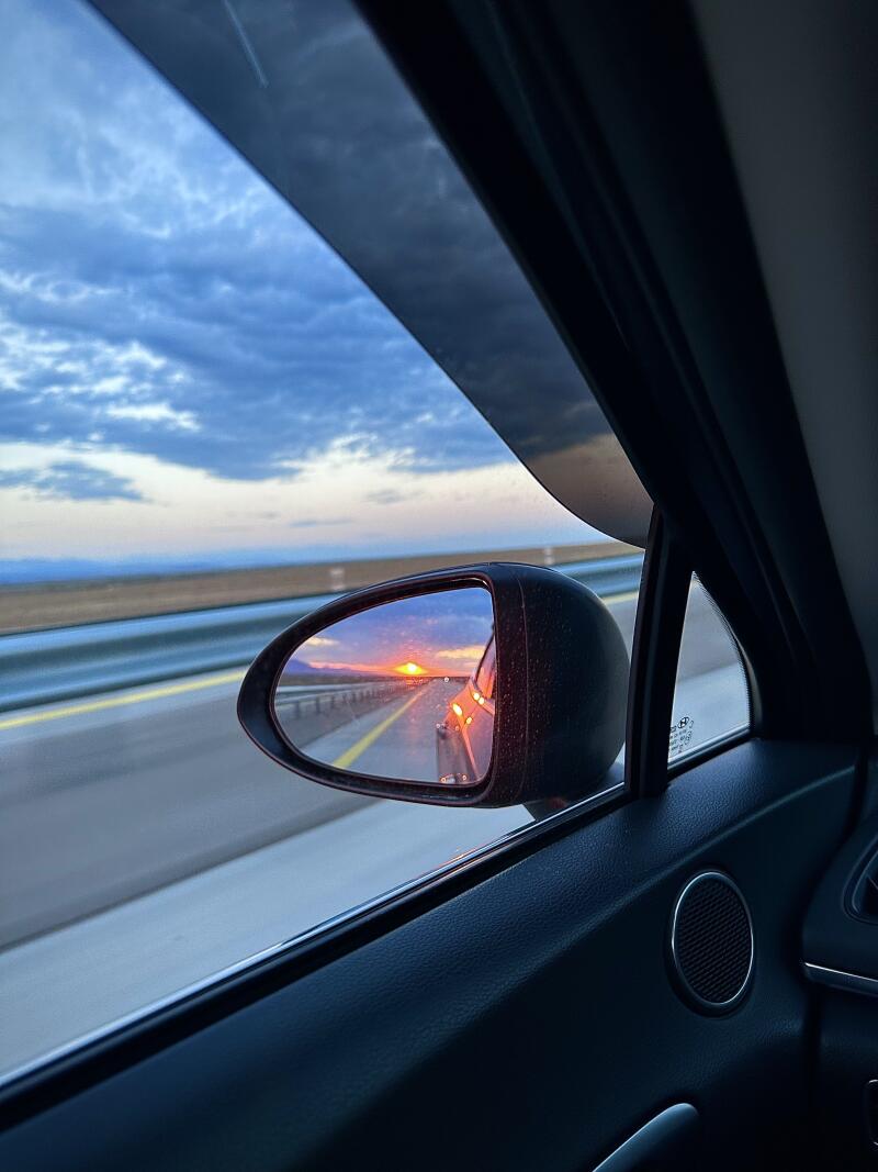 A view from inside a car shows a side mirror reflecting the sunset on a highway, with a cloudy blue sky visible through the car window.