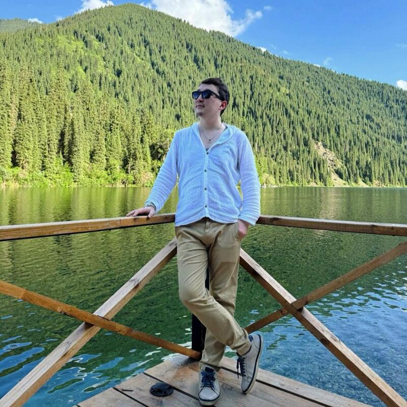 A man in sunglasses, a white shirt, and khaki pants stands on a wooden dock with a lake and tree-covered mountain in the background.