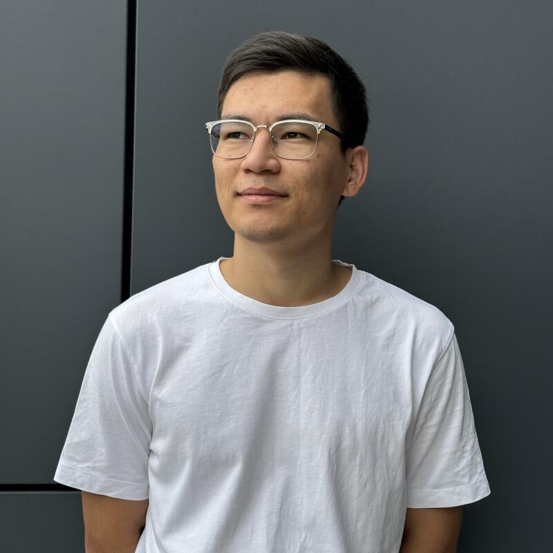 A man with glasses and a white T-shirt stands in front of a gray wall.