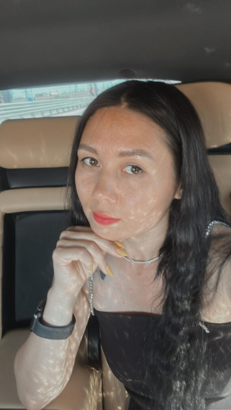 A woman with long dark hair and red lipstick is sitting in a car, resting her chin on her hand.