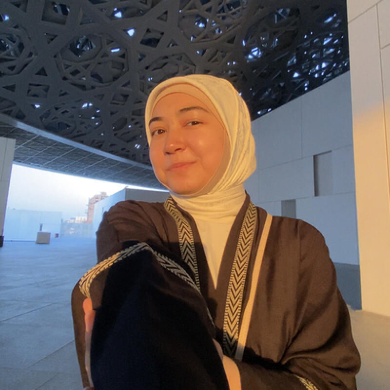 A woman wearing a white hijab and a brown abaya with patterned trim poses under an architectural structure with a geometric design.