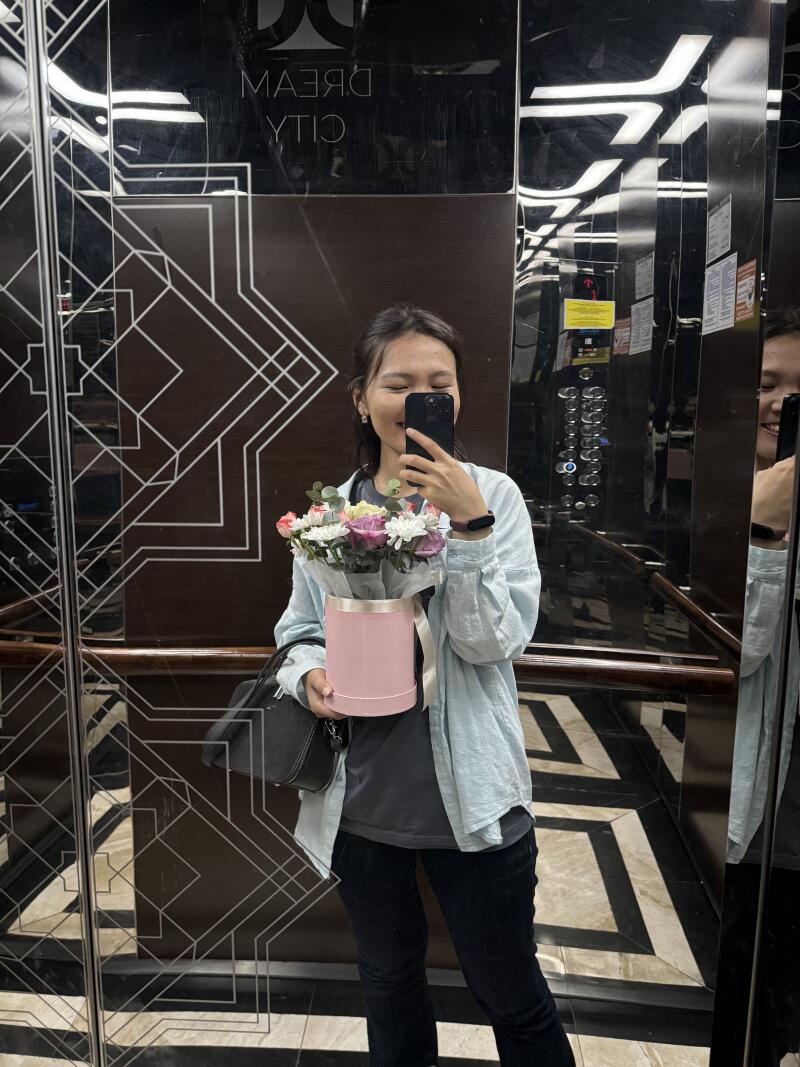 A woman is taking a selfie in an elevator, holding a bouquet of flowers in a pink box. The elevator's interior features a geometric design and the text "DREAM CITY" in reverse.