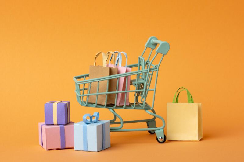 A miniature turquoise shopping cart is filled with shopping bags, with additional gift boxes and a shopping bag arranged around it against an orange background.