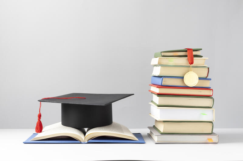 A graduation cap sits on an open book next to a stack of books with a gold medal draped over the top, all against a gray background.