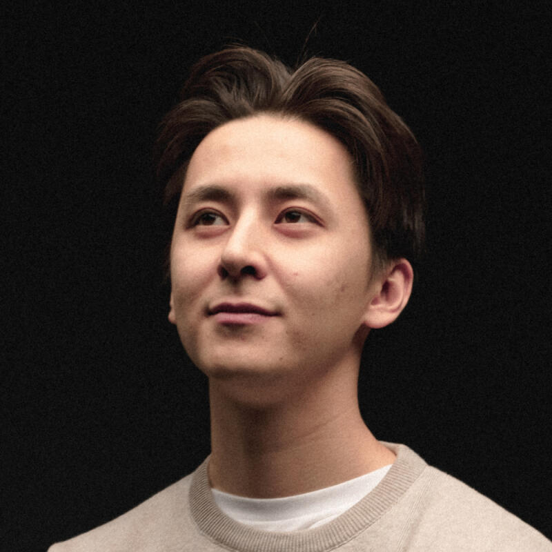 A portrait of a man with dark hair, wearing a beige sweater, looking upwards against a black background.