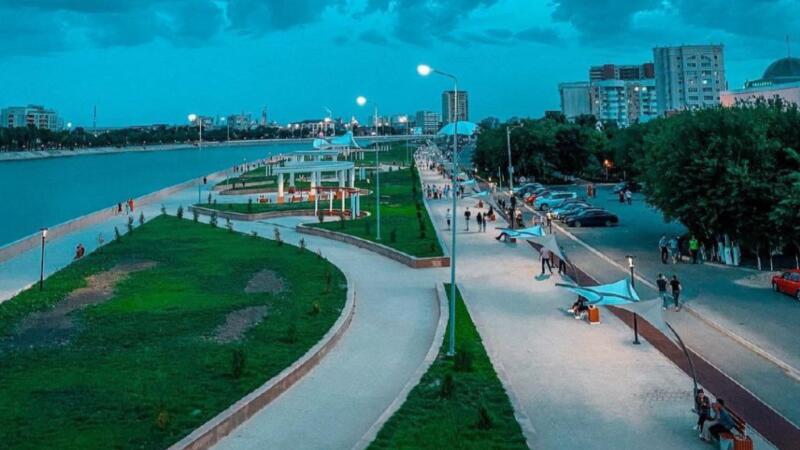 Живописный вид городского парка вдоль берега реки с пешеходными дорожками, зелеными лужайками и уличными фонарями под облачным небом.