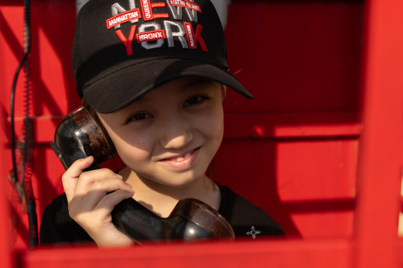 A young girl in a black cap with "NEW YORK" embroidered on it is holding a vintage black telephone receiver inside a red telephone booth, smiling slightly.