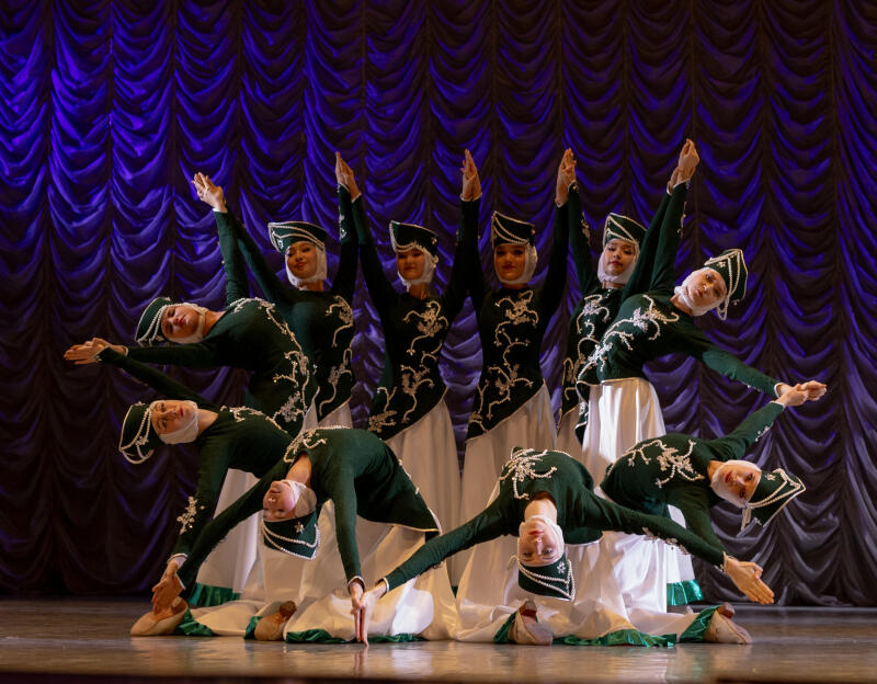 A group of women in green and white costumes are performing a dance on a stage with a blue curtain backdrop. They are arranged in a formation, some with arms raised and others bent over.