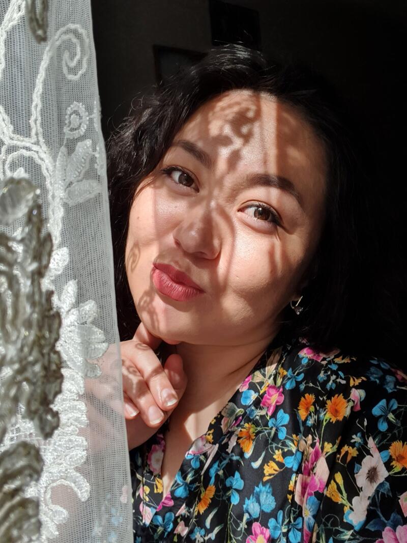 A woman poses in front of a window, with sunlight casting shadows on her face, wearing a floral shirt and holding her hand to her chin.