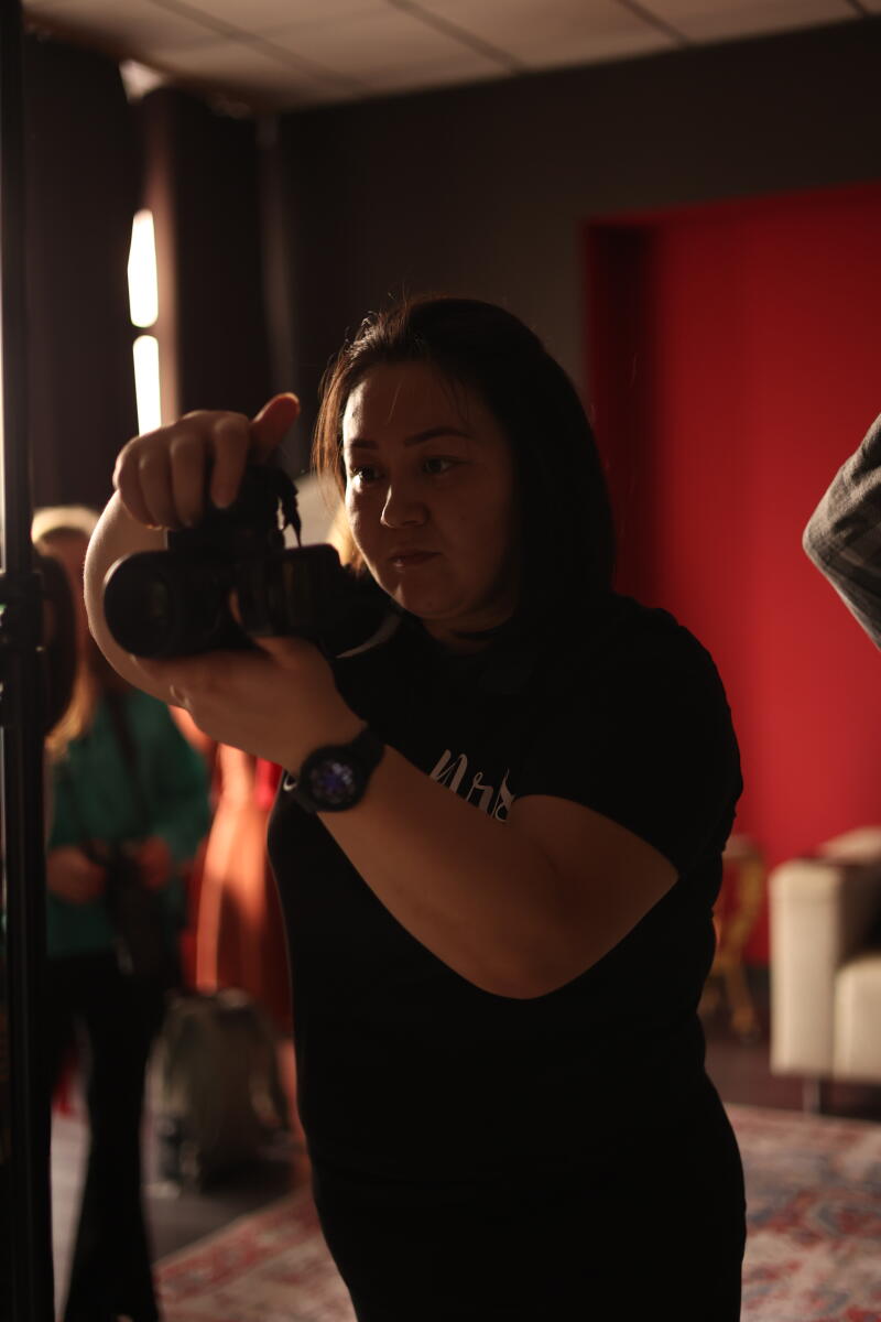 A woman in a black T-shirt is holding a camera and taking a photo in a studio setting with other people in the background.