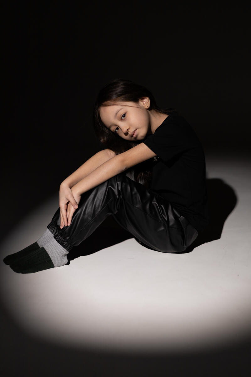 A young girl with long brown hair sits on the floor in a spotlight, wearing a black t-shirt, black leather pants, and gray and dark green socks.