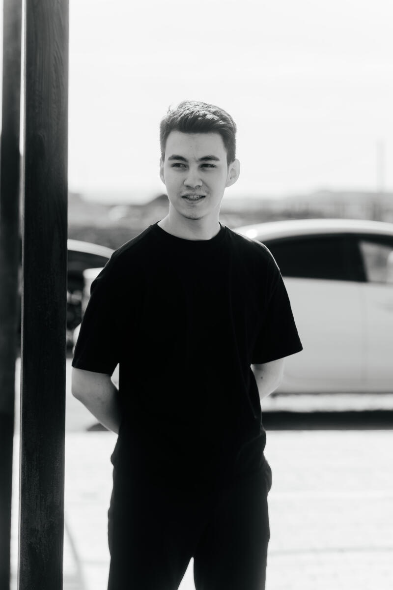 A black and white shot features a young man in a dark T-shirt and pants, with his hands clasped behind his back, standing next to a dark post and a blurred white car.