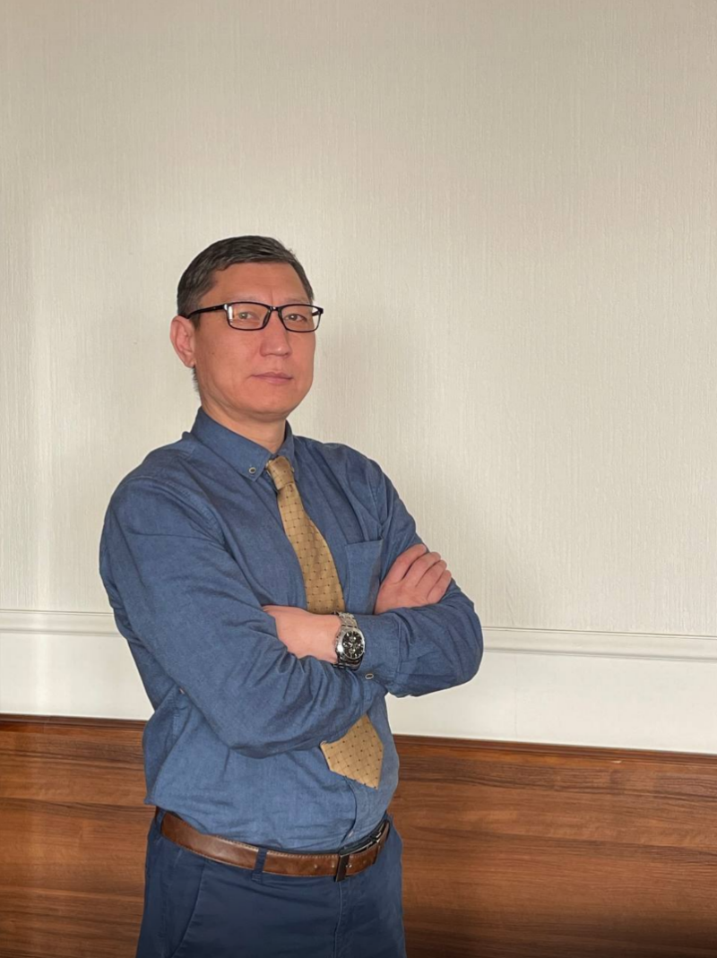 A man with glasses, wearing a blue shirt and a gold tie, stands with his arms crossed.