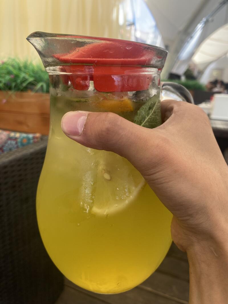 A hand holds a glass pitcher filled with yellow lemonade, containing sliced citrus and mint leaves, and sealed with a red lid.