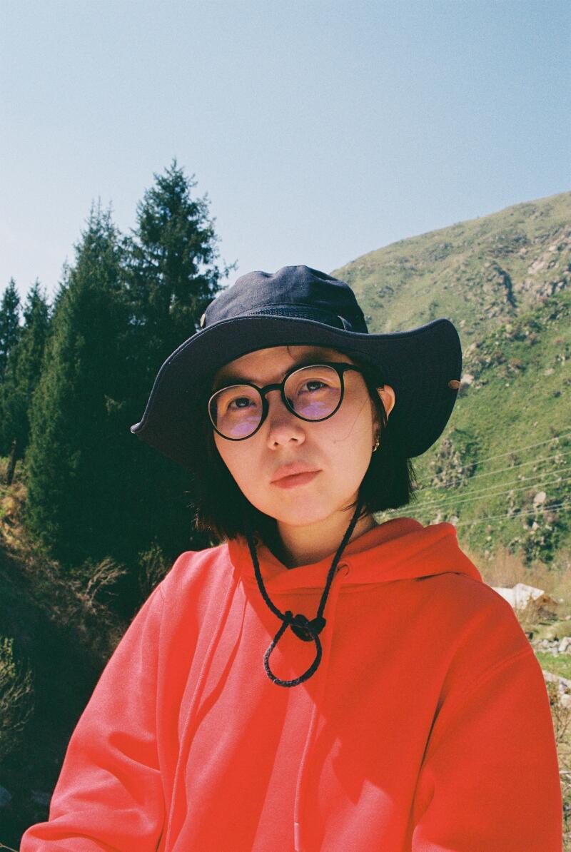 A person wearing glasses and a dark hat is wearing a red hoodie, standing outside with trees and a hill in the background.