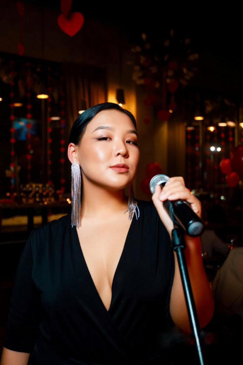 A woman in a black dress and long, silver earrings holds a microphone, singing in a room decorated with red hearts.