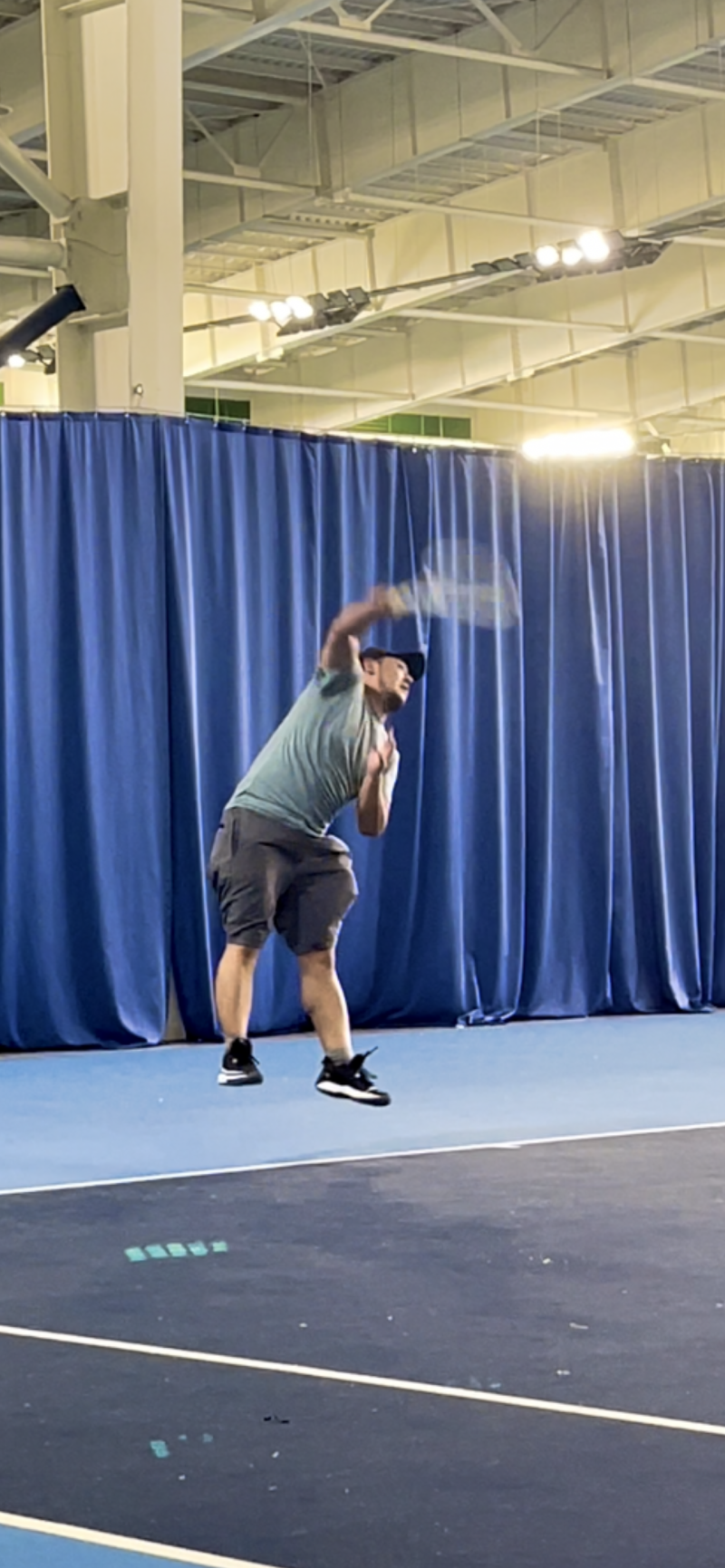 A man is playing tennis, swinging his racket in an indoor court with blue curtains in the background.