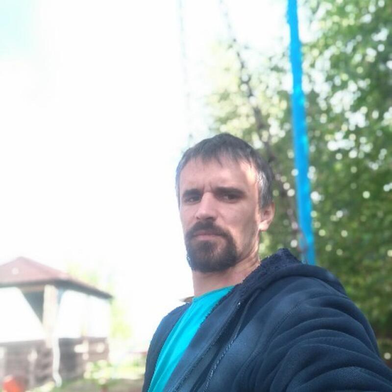 A man with a beard and a blue shirt under a blue jacket takes a selfie outdoors.