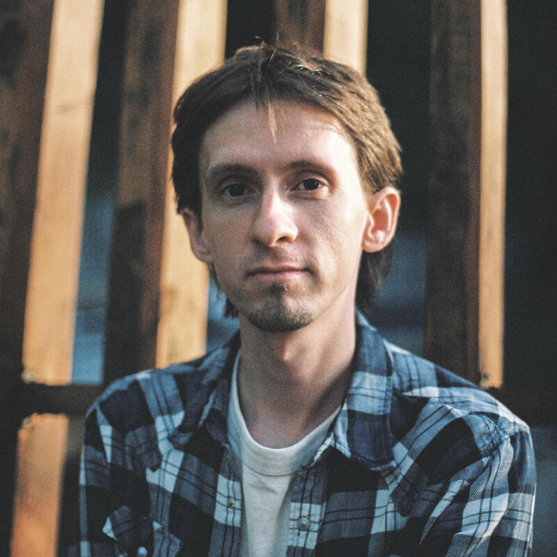 A man with short brown hair and a slight beard is wearing a blue and gray plaid shirt over a white t-shirt, standing in front of a wooden structure.