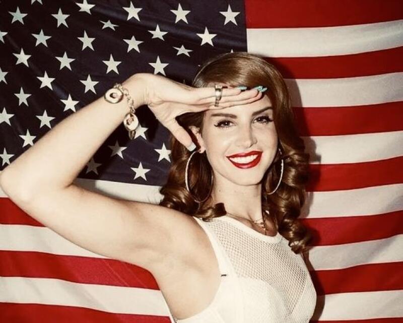 A woman with brown hair and red lipstick salutes in front of an American flag.