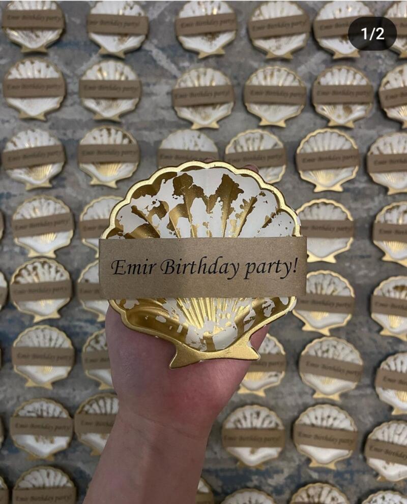 The image shows a hand holding a decorative seashell with a paper label that reads "Emir Birthday party!", surrounded by many similar seashell favors.