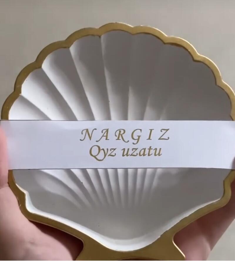 A white, shell-shaped dish with a gold rim is held, displaying the text 'NARGIZ Qyz uzatu' in gold lettering.