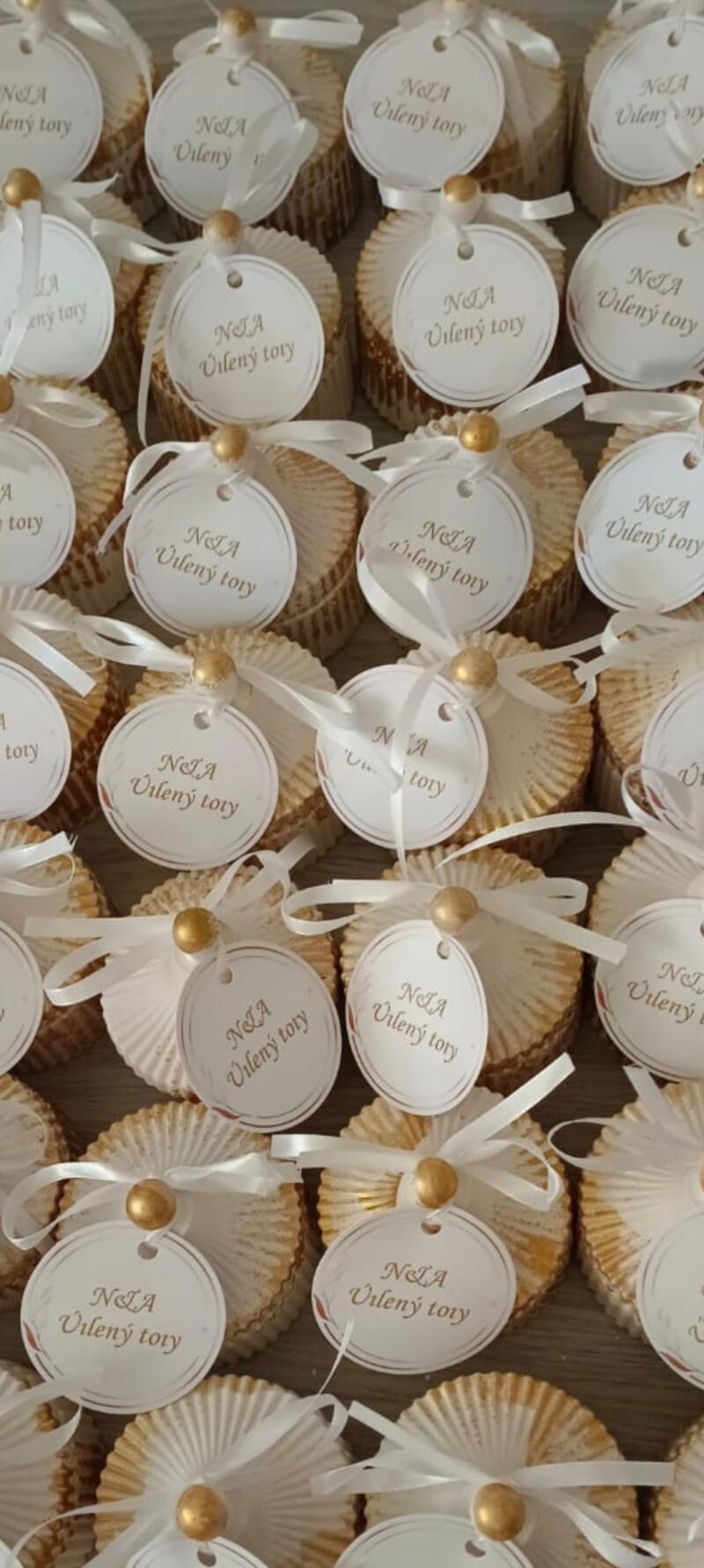 The image shows numerous cupcakes in white and gold wrappers, each adorned with a white tag that reads "Nela Vileny tory" and tied with a white ribbon and a gold bead.