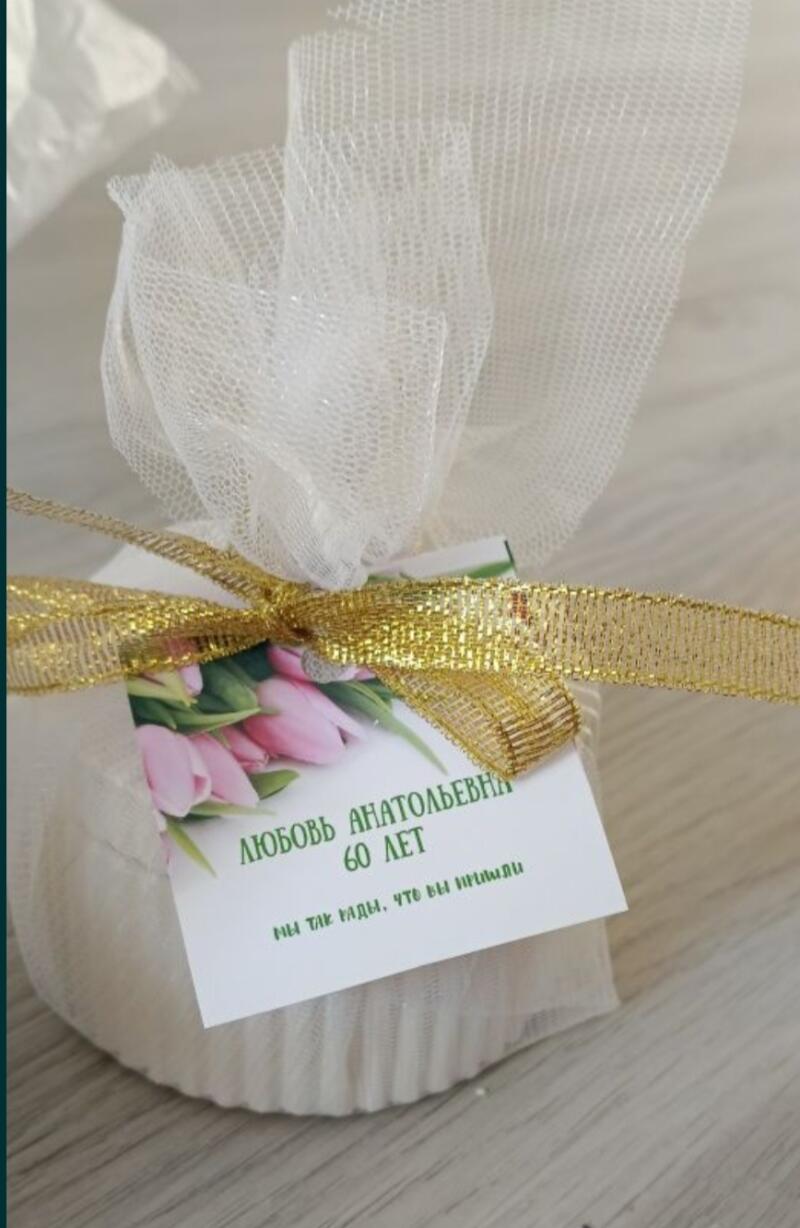 A gift wrapped in white mesh fabric and tied with a gold ribbon, with a card that reads "ЛЮБОВЬ АНАТОЛЬЕВНА 60 ЛЕТ. МЫ ТАК РАДЫ, ЧТО ВЫ ПРИШЛИ" in green text, and an image of pink tulips.