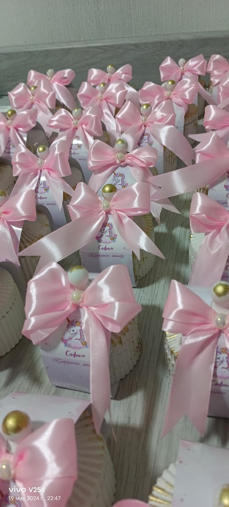 The image shows several small, decorated gift boxes, each adorned with a pink ribbon tied into a bow and a small bead; the boxes have a label featuring a unicorn and the text "Сафия қырқынан шығару".